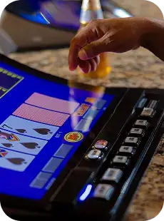 A hand hovers over the buttons of a video poker machine showing a hand of clubs. A bottle, possibly of beer, is seen in the background on a granite countertop.