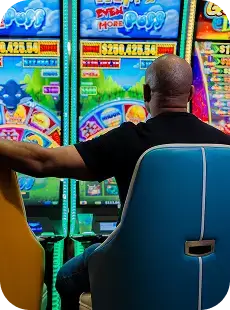A man sits alone in front of brightly lit slot machines at a casino, his arm resting on an empty chair beside him.