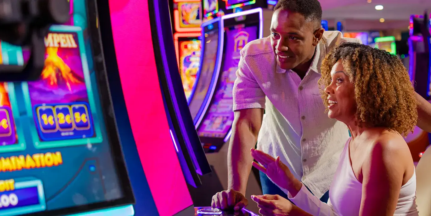 A woman and a man smile and enjoy playing a colorful slot machine in a lively casino, with bright lights and other slot machines visible in the background.