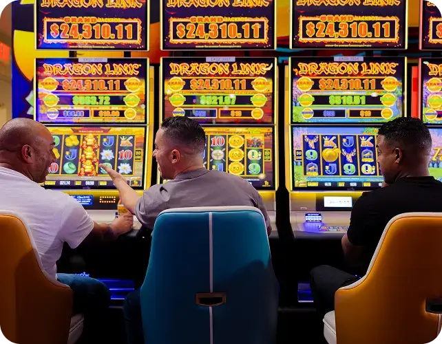 Three people sit side by side playing colorful Dragon Link slot machines at a casino, with bright digital displays showing jackpots and game graphics above them.
